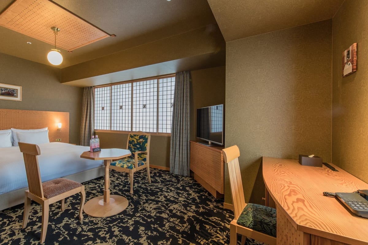 A hotel room with a bed, a desk, a chair, a TV, and a window with curtains.