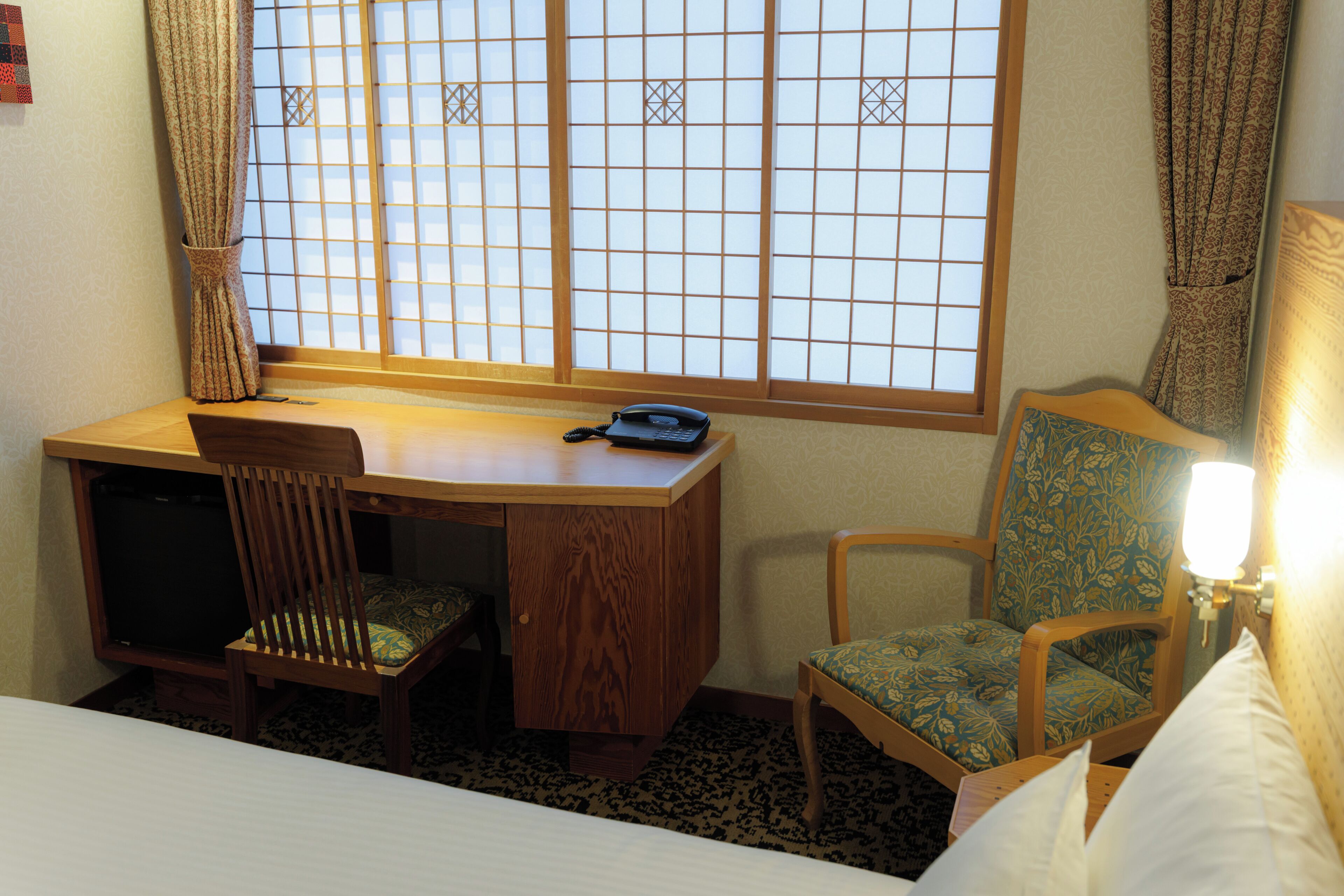 A hotel room with a wooden desk, a chair, a bed, and a window with curtains.