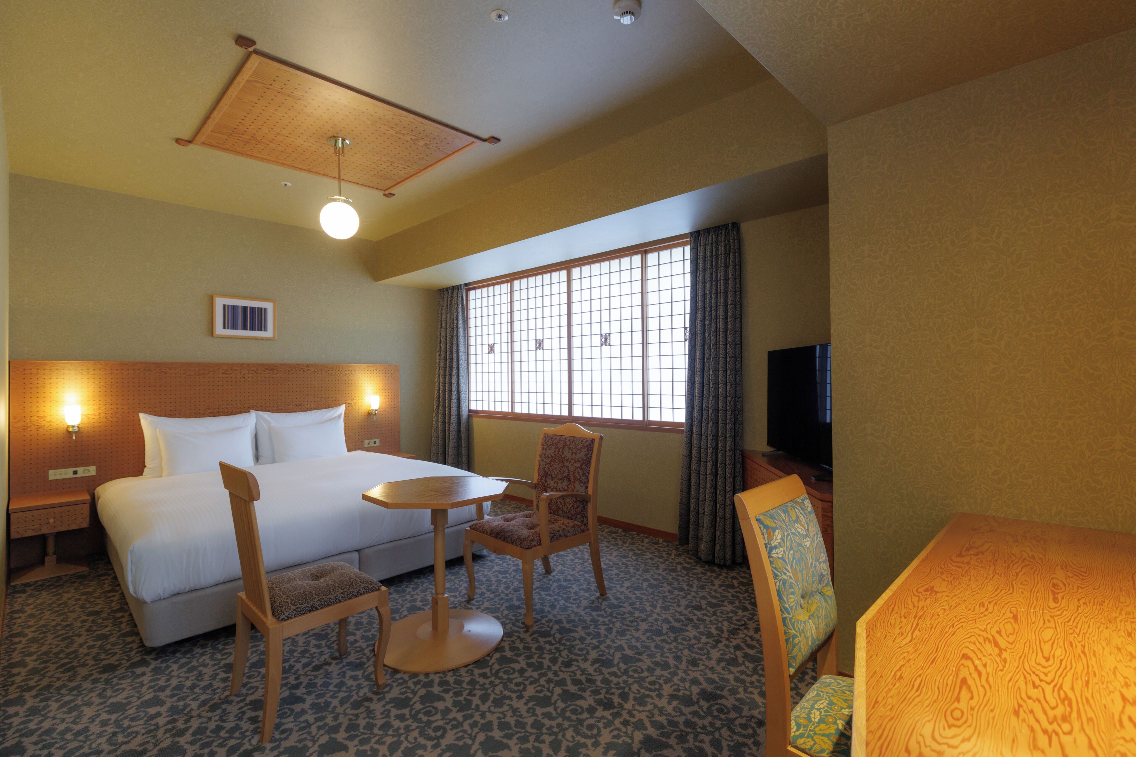 A hotel room with a bed, a desk, two chairs, a television, and a window with curtains.