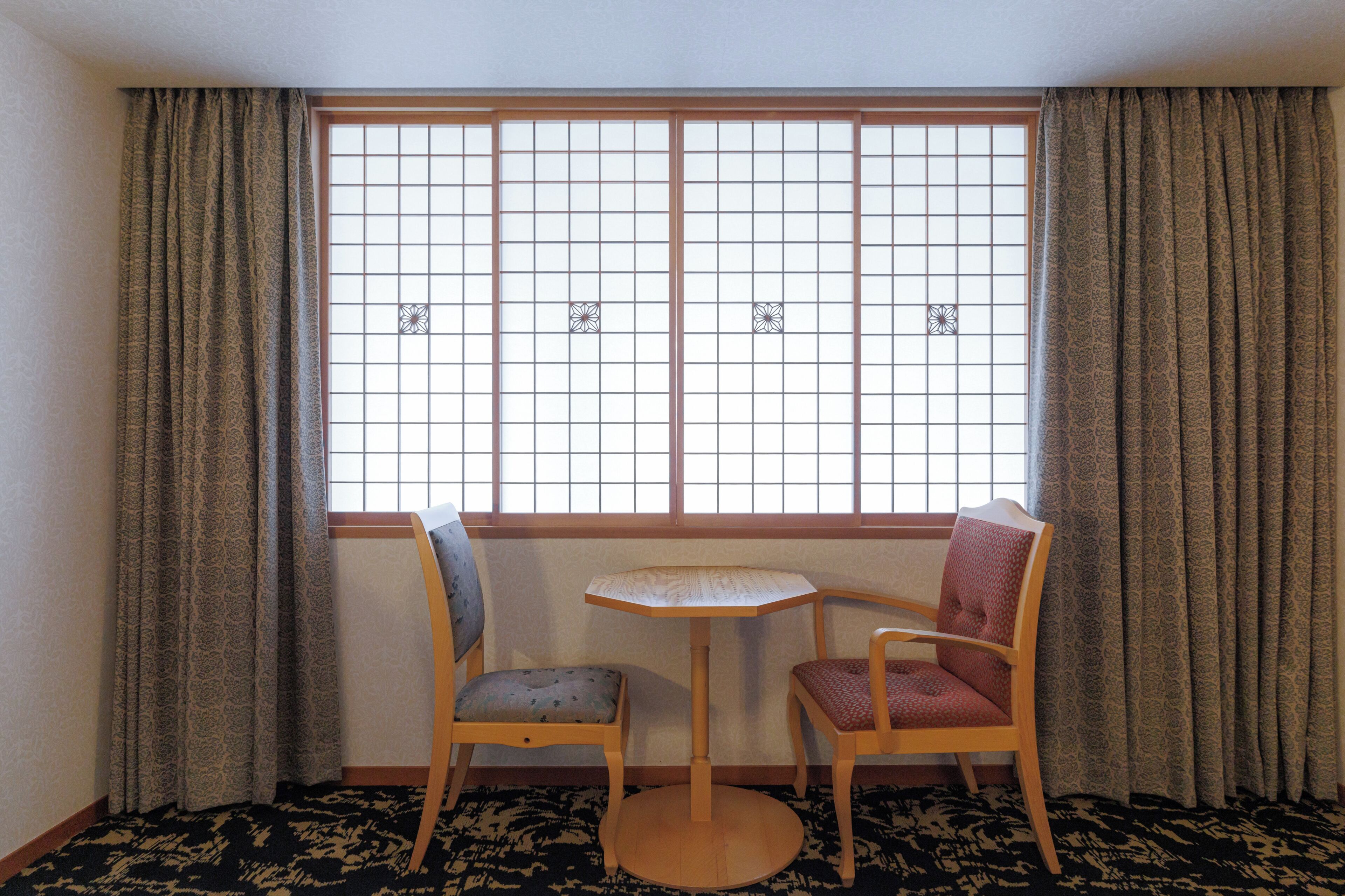 Une pièce avec un sol recouvert de tatamis, une fenêtre à panneaux coulissants de style shoji, deux chaises et une petite table ronde.