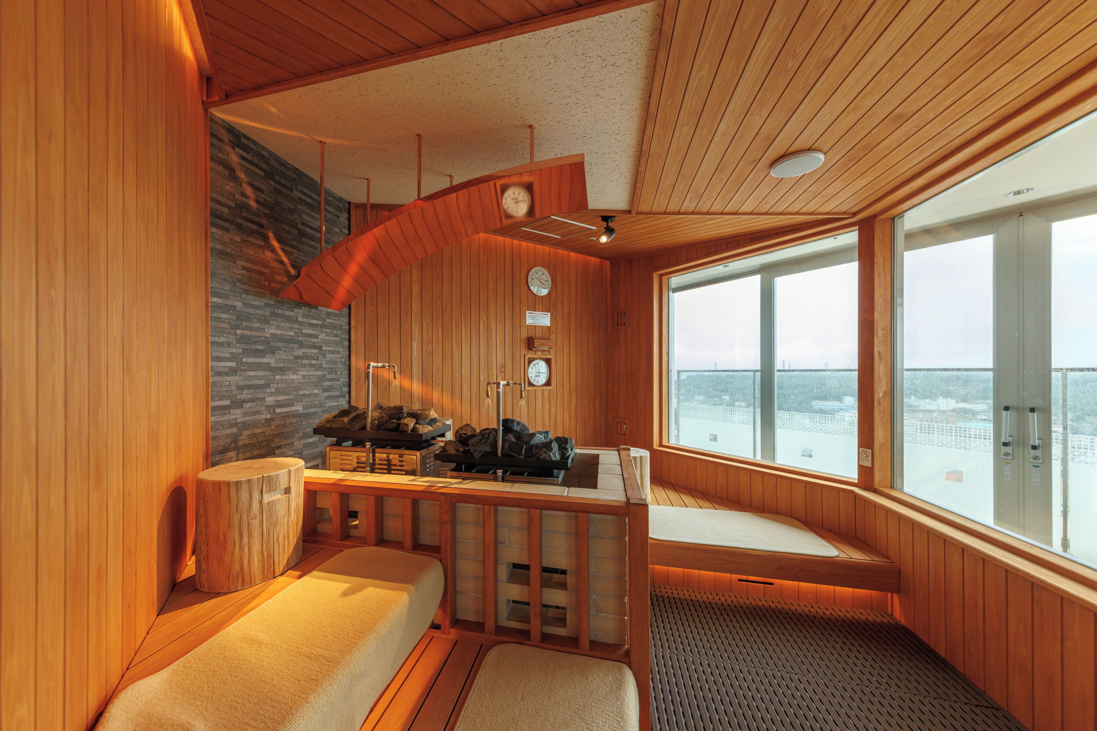 A sauna room with wooden walls, a stone wall, and a large window. There are benches and a table with towels.