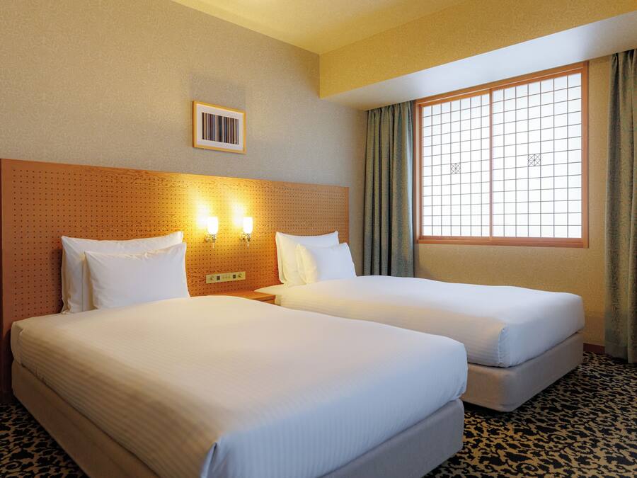 A hotel room with two beds, a window with curtains, and a wall-mounted light fixture.