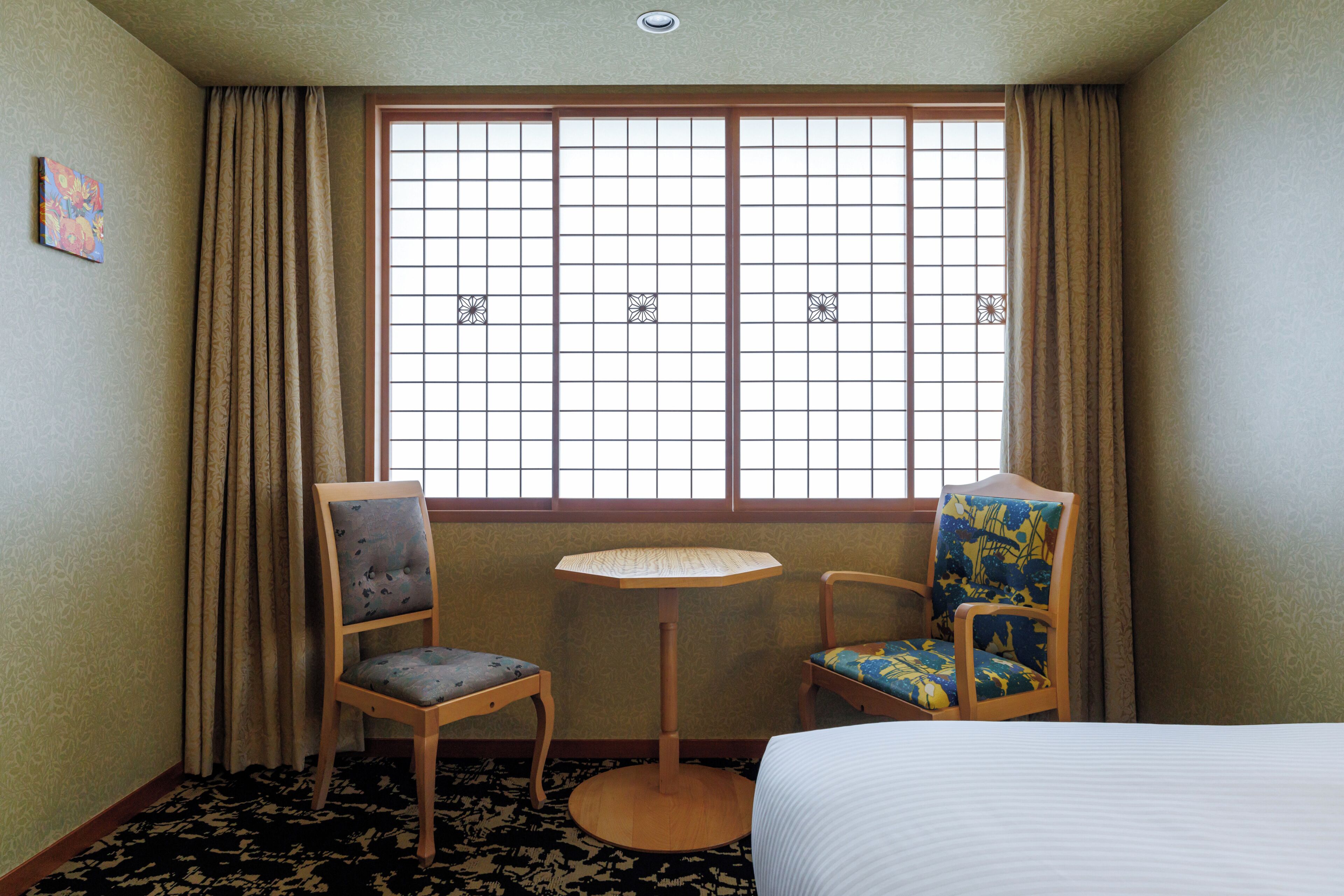 Ein Hotelzimmer mit einem Bett, zwei Sesseln, einem kleinen Tisch und einem Fenster mit Rasteroptik.
