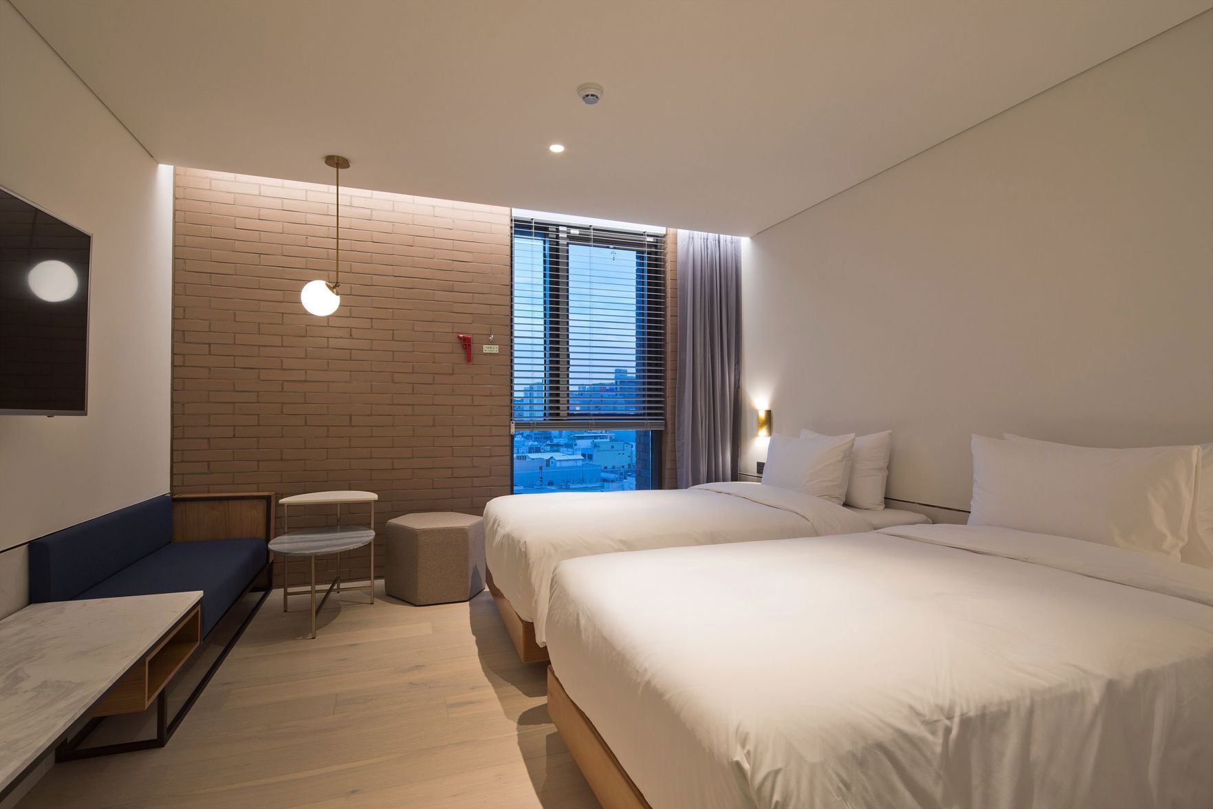 Een moderne hotelkamer met een accentmuur van baksteen, een groot raam met zonwering, een bank, een klein rond tafeltje en een bed met witte lakens.