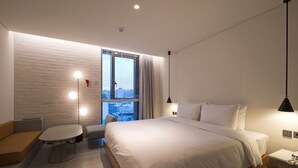 A modern hotel room with a large bed, a small round table, a sofa, and a window with blinds.