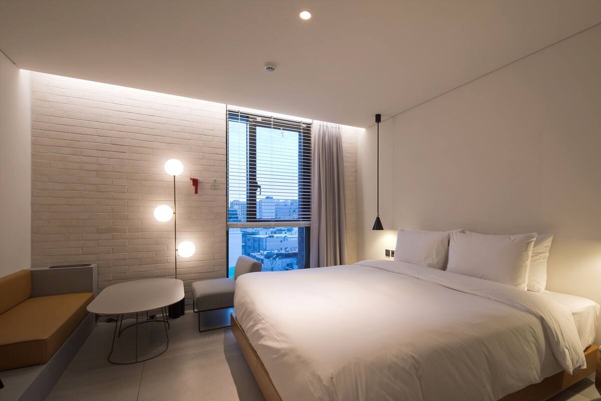 A modern hotel room with a large bed, a small round table, a sofa, and a window with blinds.