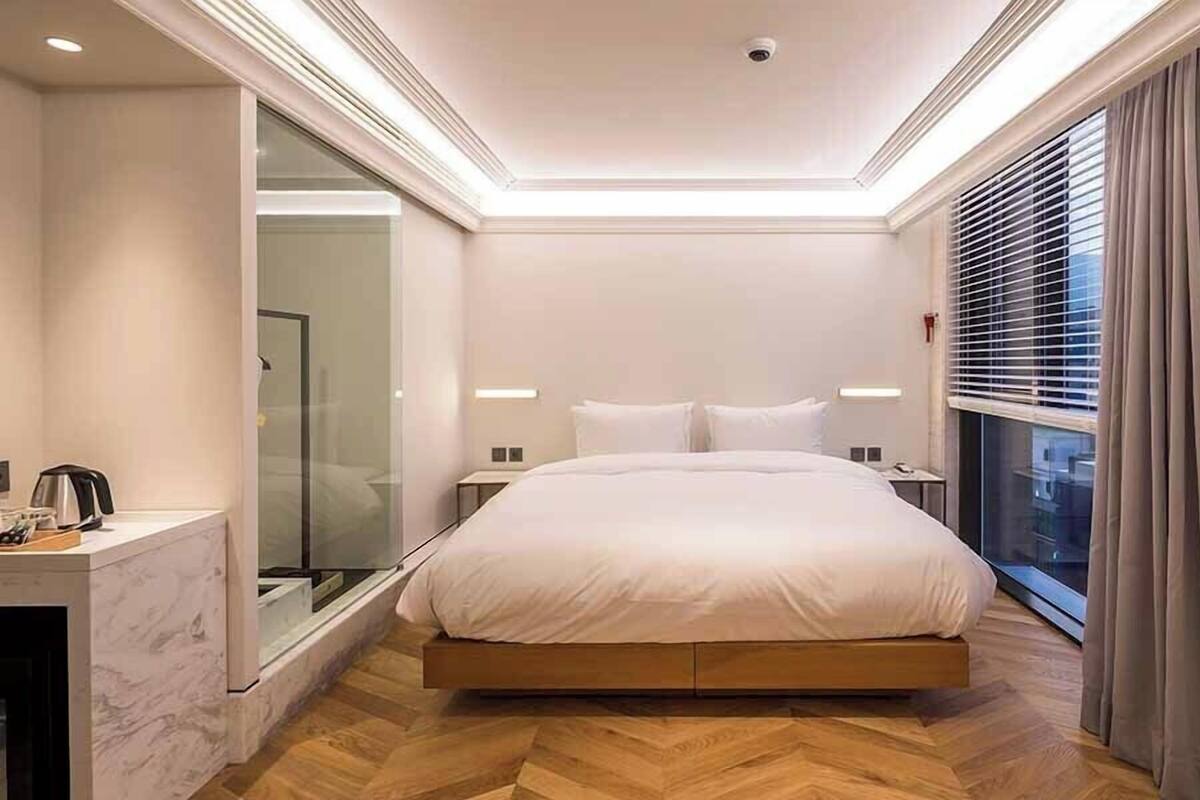 A modern hotel room with a large bed, a glass shower enclosure, and a view of the city through the window.