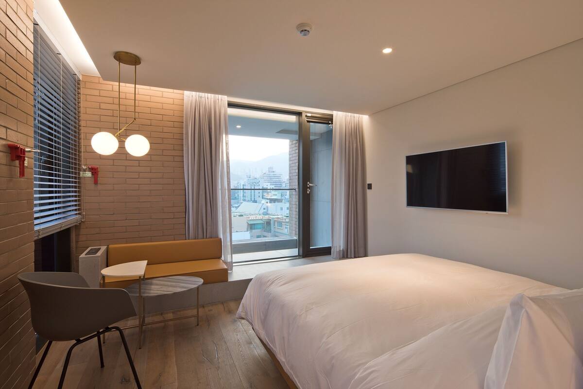 A modern hotel room with a bed, a TV, a seating area, and a balcony with a city view.