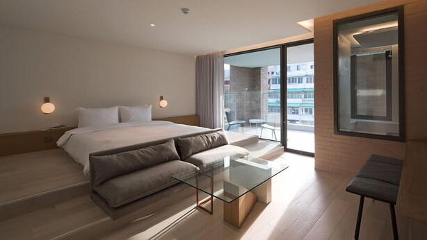 A modern hotel room with a large bed, a glass coffee table, a sofa, and a balcony with a view of the city.