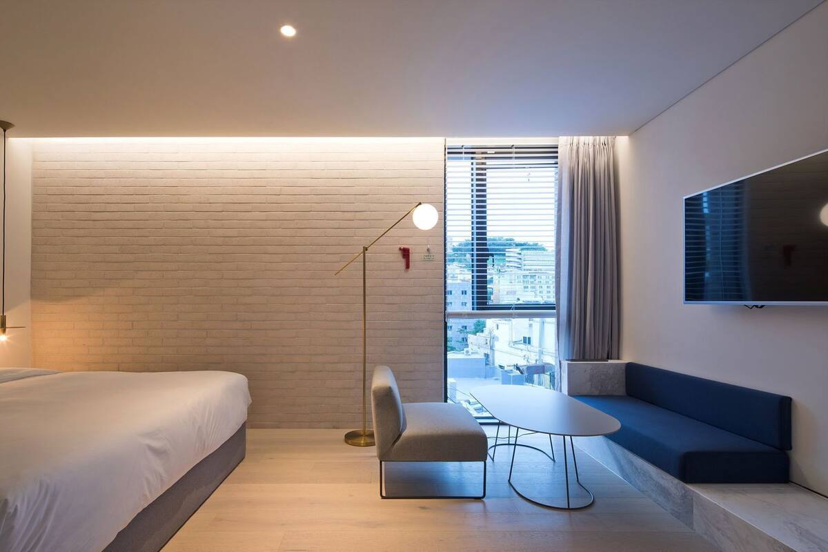 A modern hotel room with a bed, a desk, a chair, a TV, and a window with blinds.