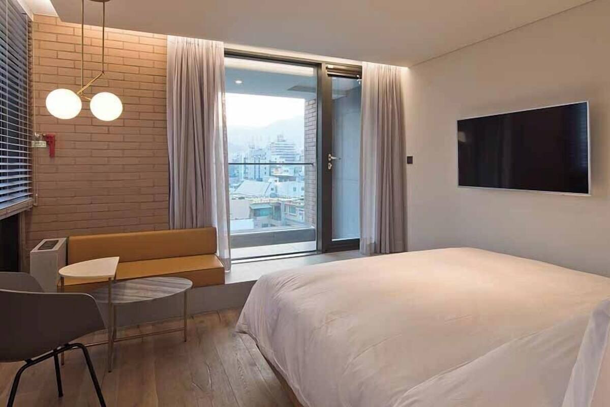 A modern hotel room with a bed, a sofa, a TV, and a balcony with a city view.