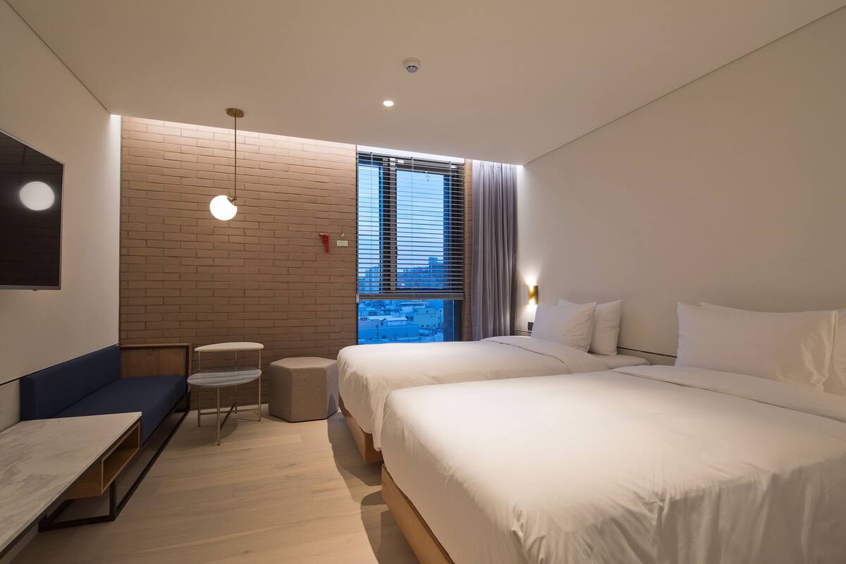 A modern hotel room with a large bed, a seating area with a table, and a view of the city through a window.