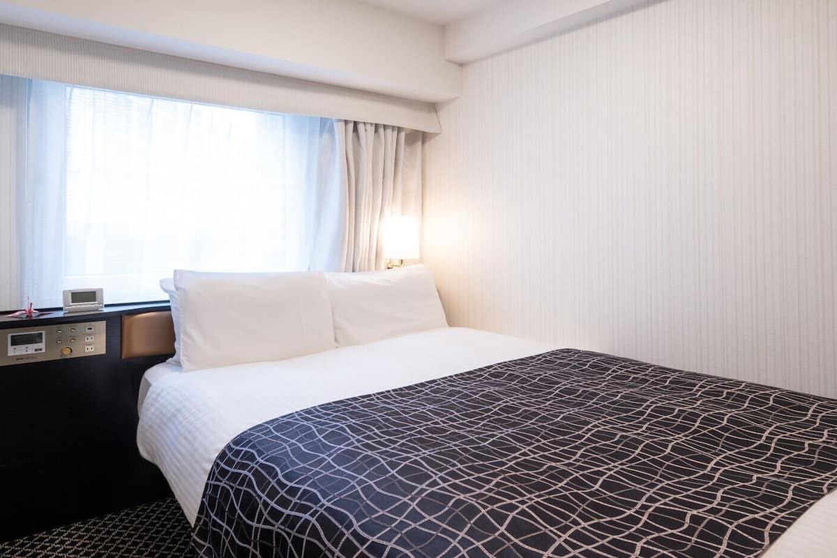 Une chambre d’hôtel avec un lit, un panneau de commande, une fenêtre avec des rideaux et un mur orné de lignes verticales.