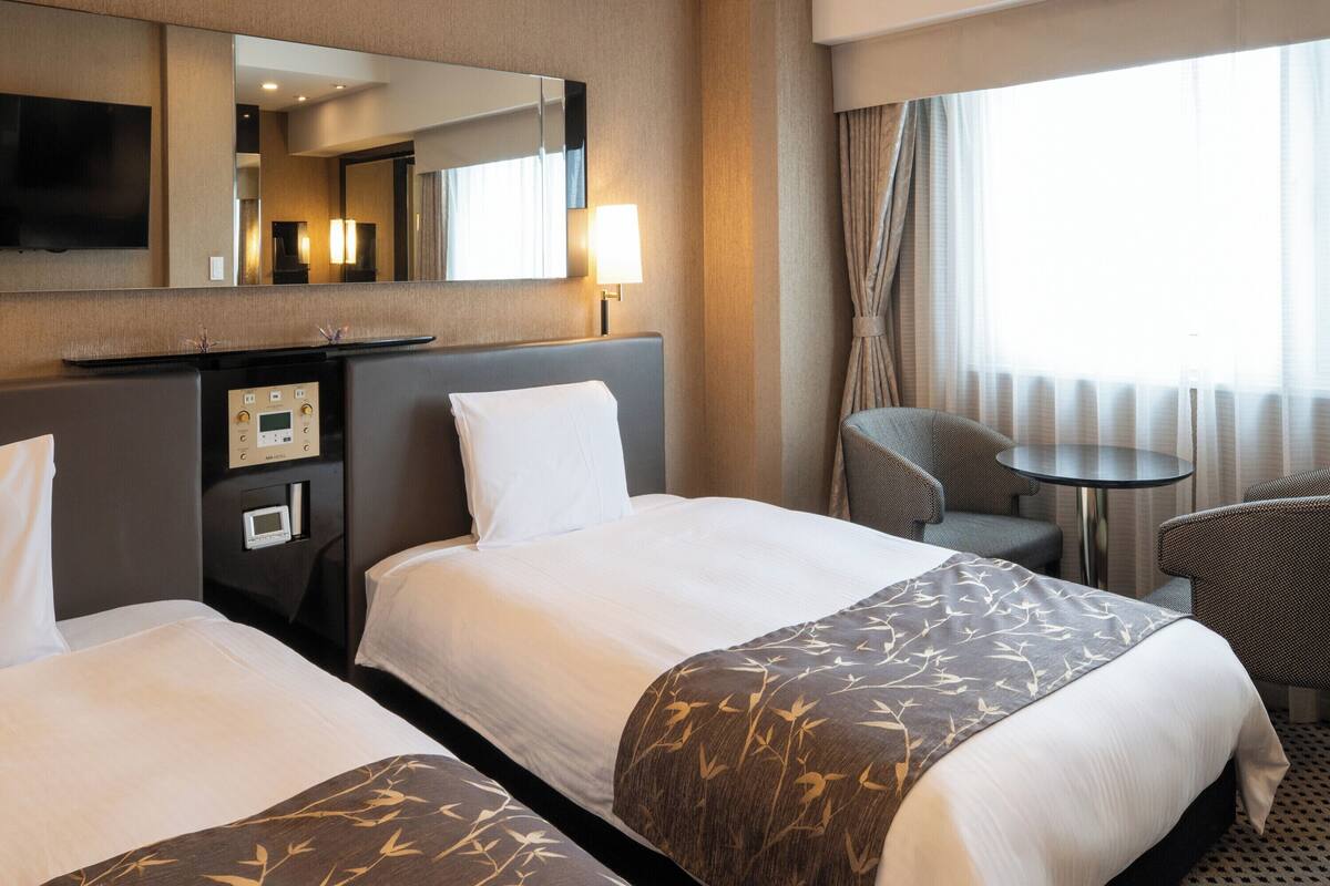 Une chambre d’hôtel avec deux lits, un bureau, une chaise, une petite table, un téléviseur et une fenêtre avec des rideaux.