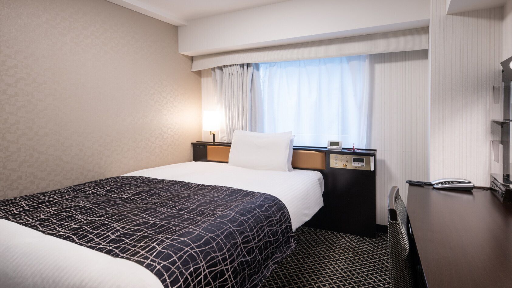 Una habitación de hotel con cama, mesita de noche con lámpara, teléfono y ventana con cortinas.