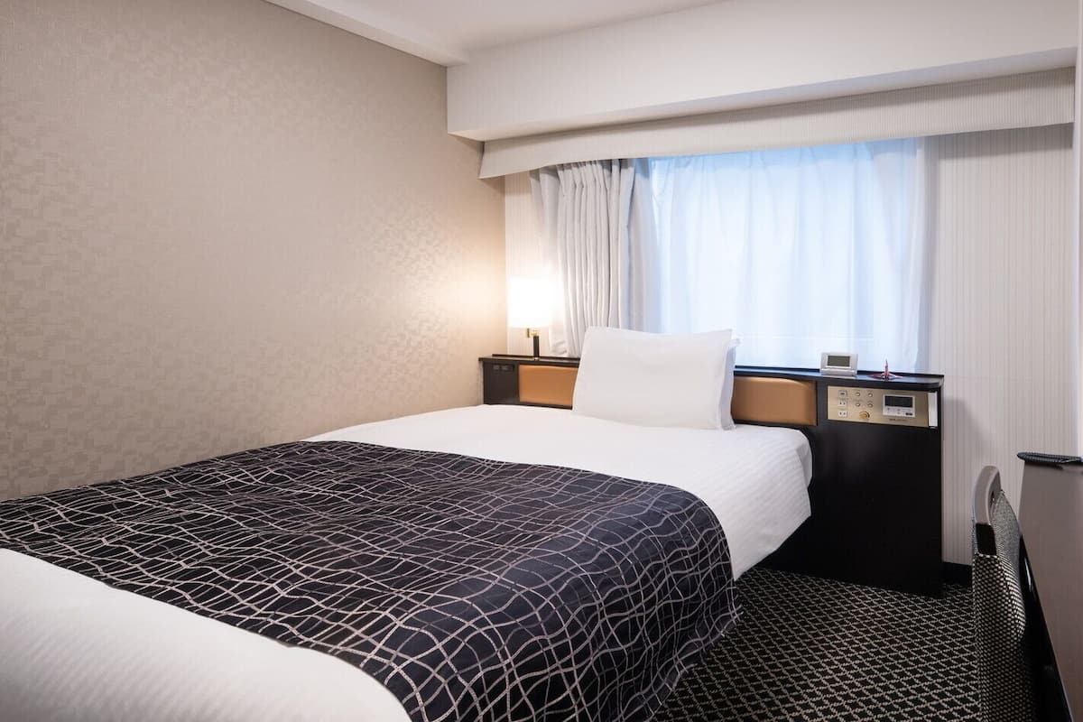 A hotel room with a bed, a bedside table with a lamp, a phone, and a window with curtains.