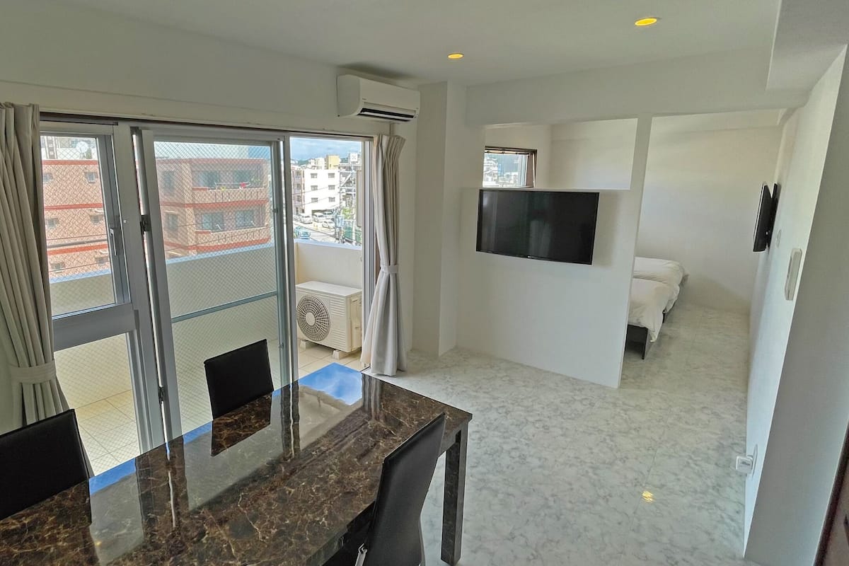 A hotel room with a dining area, a TV mounted on the wall, and a balcony with a view of the city.