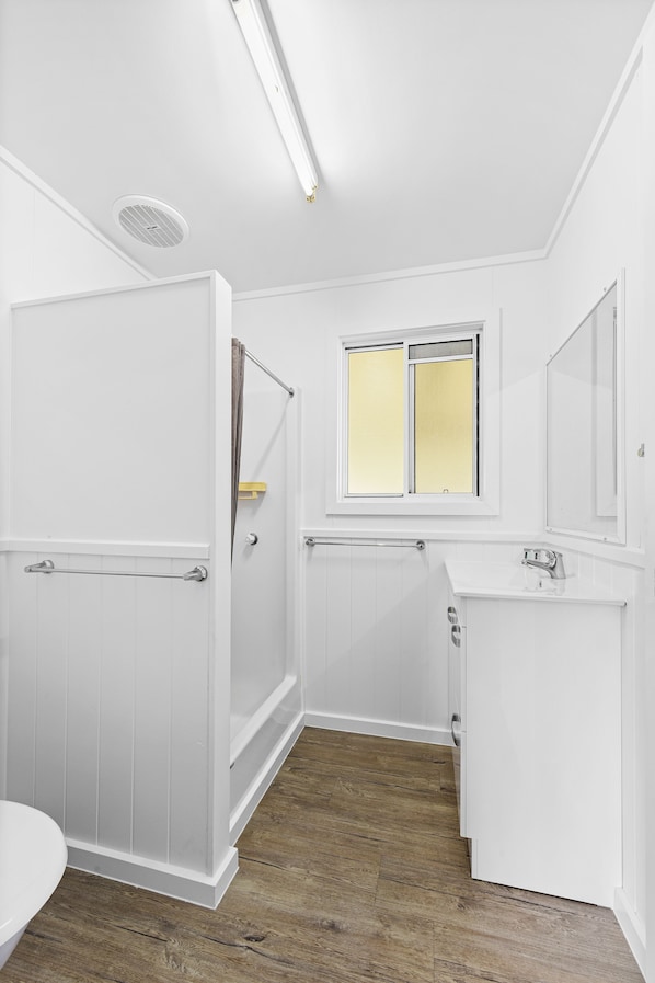 A bathroom with a shower, toilet, and sink, featuring white walls and wooden flooring.