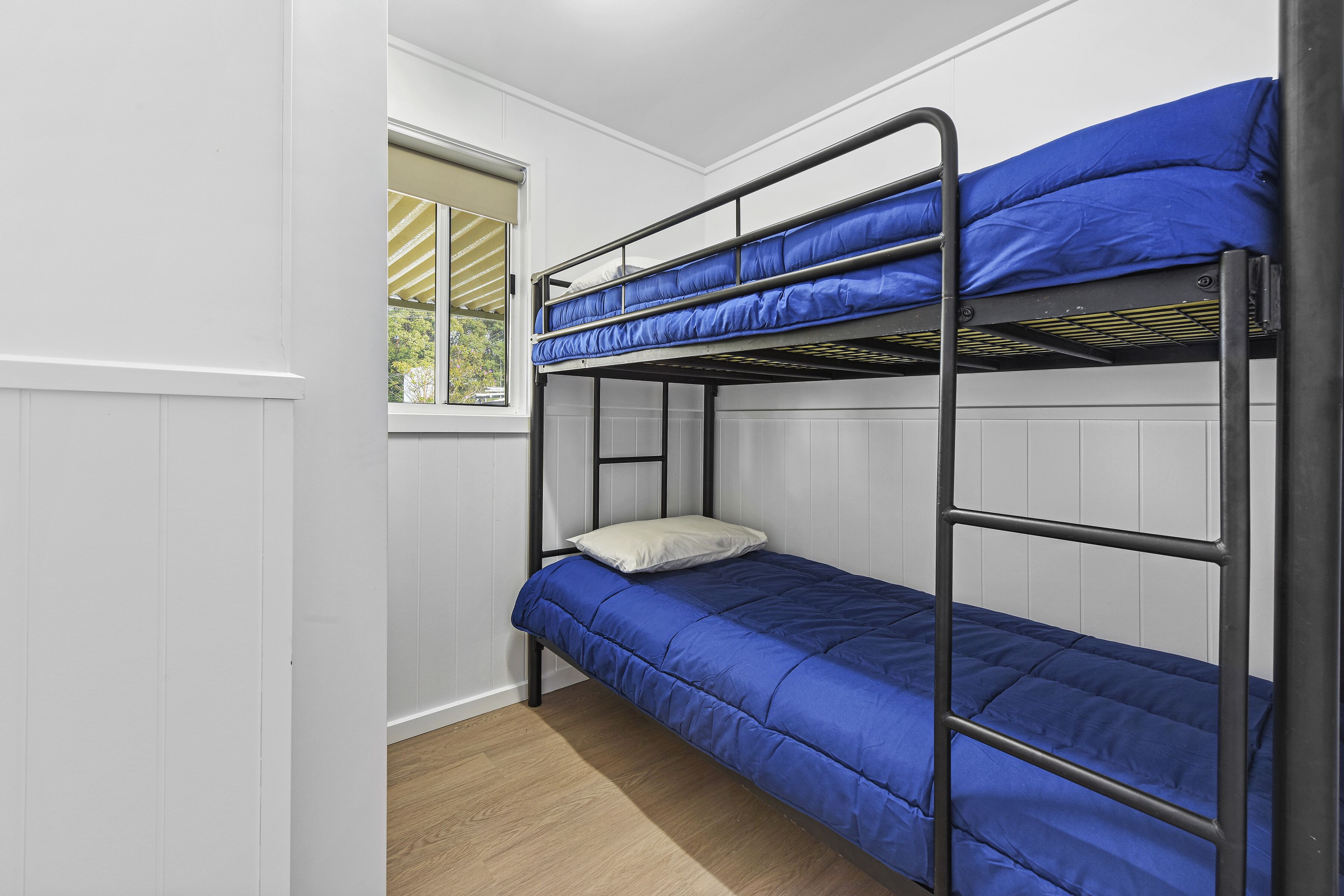Une chambre avec un lit superposé à deux niveaux, des murs blancs, une fenêtre avec des stores et un plancher en bois.