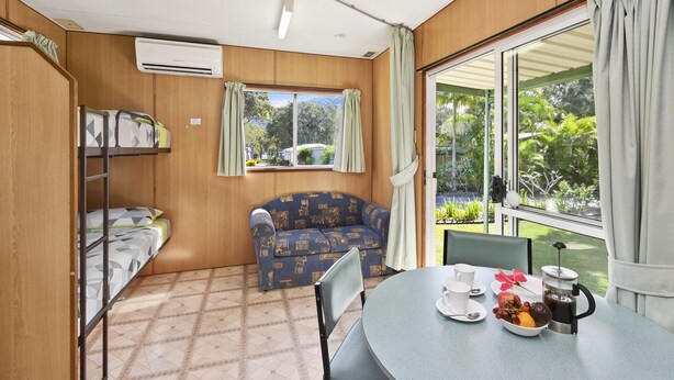A room with bunk beds, a sofa, a dining table with breakfast items, and a large window with a view of greenery. - Diamond Waters Caravan Park (Dunbogan)