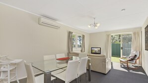 A modern living room with a glass dining table, white chairs, a beige sofa, and a television. - Diamond Waters Caravan Park (Dunbogan)