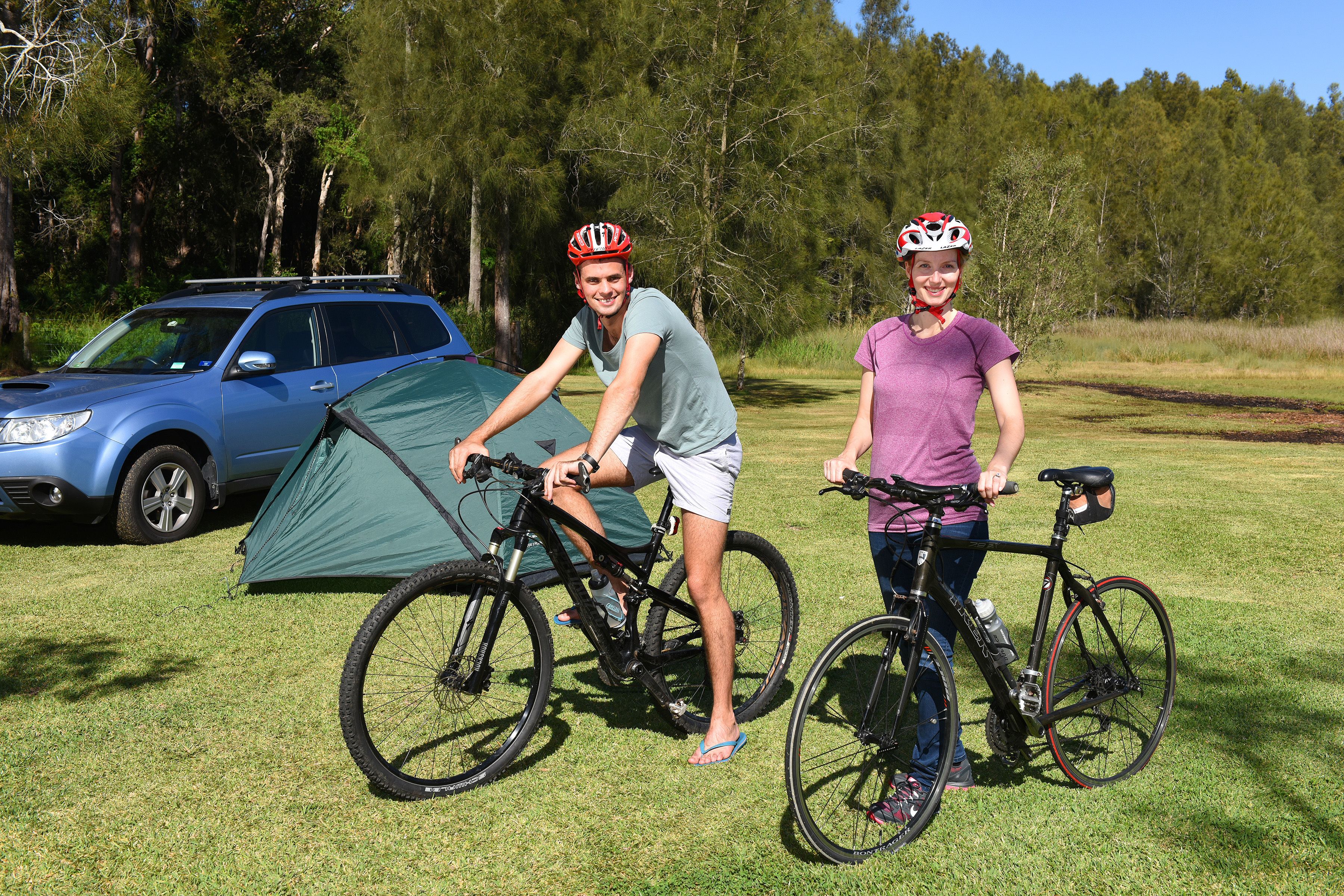 Two people on bicycles near a tent and a car in a grassy area with trees in the background.
