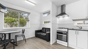 A modern kitchen with white cabinetry, a black stove, and a dining area with a black leather sofa and chairs. - Diamond Waters Caravan Park (Dunbogan)