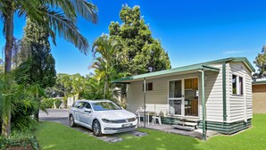 A white car parked in front of a small, single-story house with a green roof, surrounded by lush greenery and a clear blue sky. - Diamond Waters Caravan Park (Dunbogan)