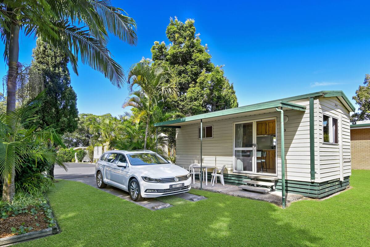 A white car parked in front of a small, single-story house with a green roof, surrounded by lush greenery and a clear blue sky.