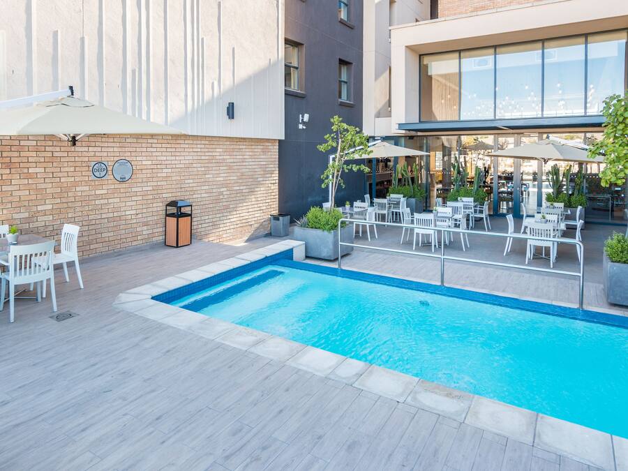 Un área de piscina exterior moderna con un muro de ladrillo, mobiliario exterior blanco y un porche con paredes de vidrio.