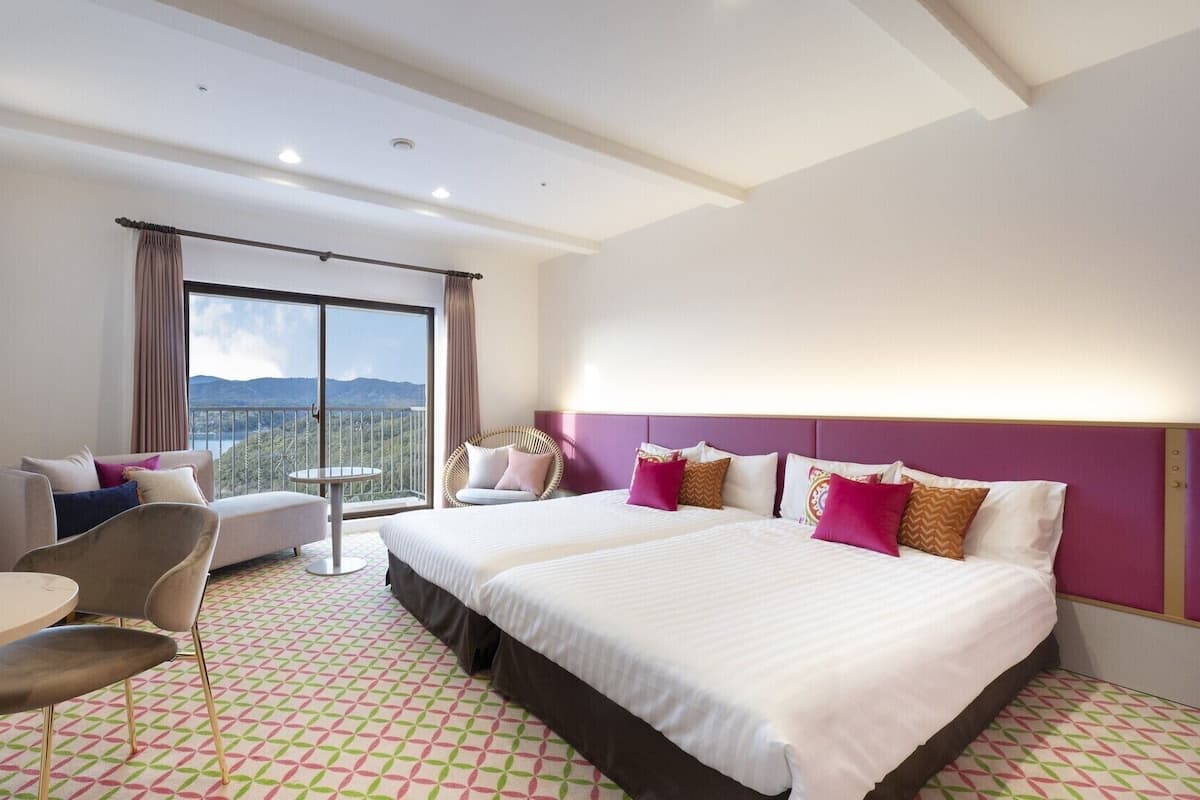 A hotel room with a large bed, a sofa, a chair, a small table, and a balcony with a view.