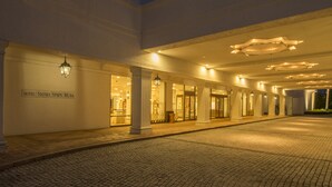 Entrada de hotel com caminho de pedras, colunas imponentes e sinalização iluminada.