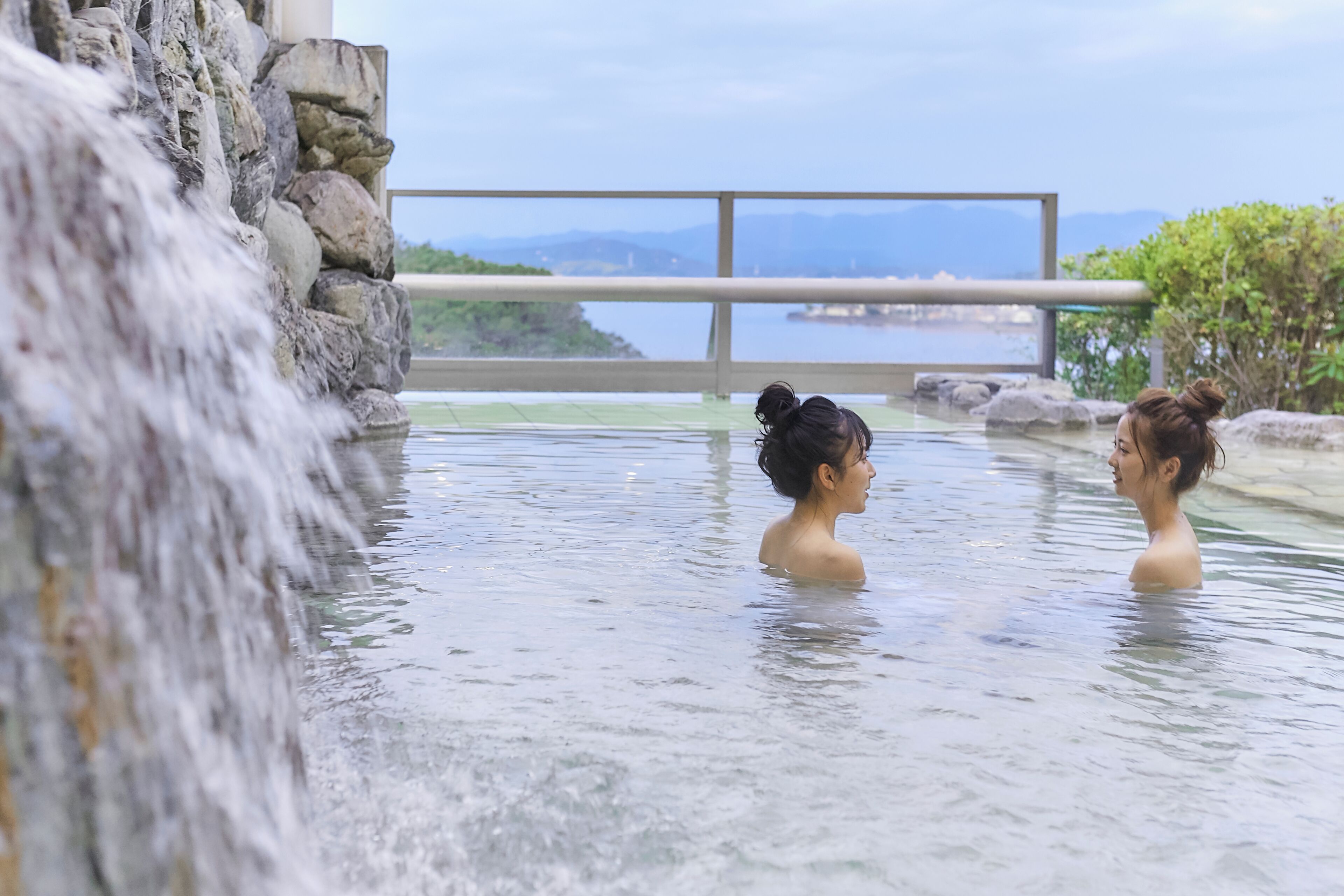 Zwei Personen in einer heißen Quelle mit Wasserfall, einer Steinmauer und Blick auf ferne Berge.