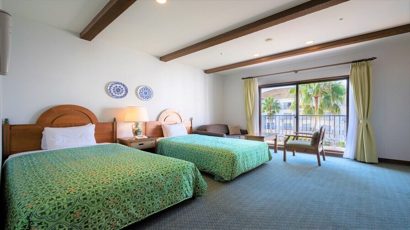Quarto de hotel com duas camas, uma cadeira, uma mesa e uma janela com cortinas.