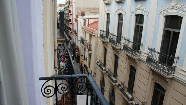 A narrow street with buildings on either side, featuring balconies with wrought iron railings and a mix of architectural styles. - Hostal Mayor (Alicante)