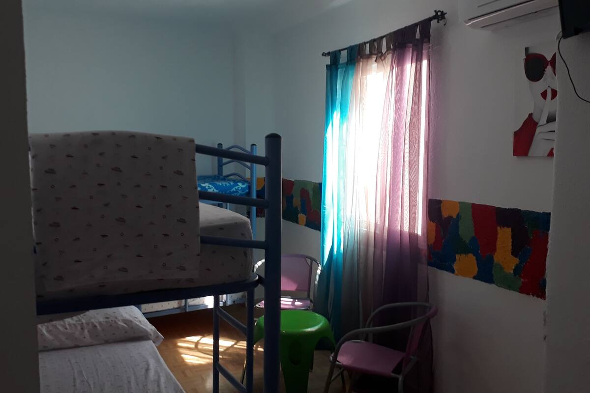 A dormitory room with bunk beds, a green plastic chair, a pink chair, and colorful wall art.