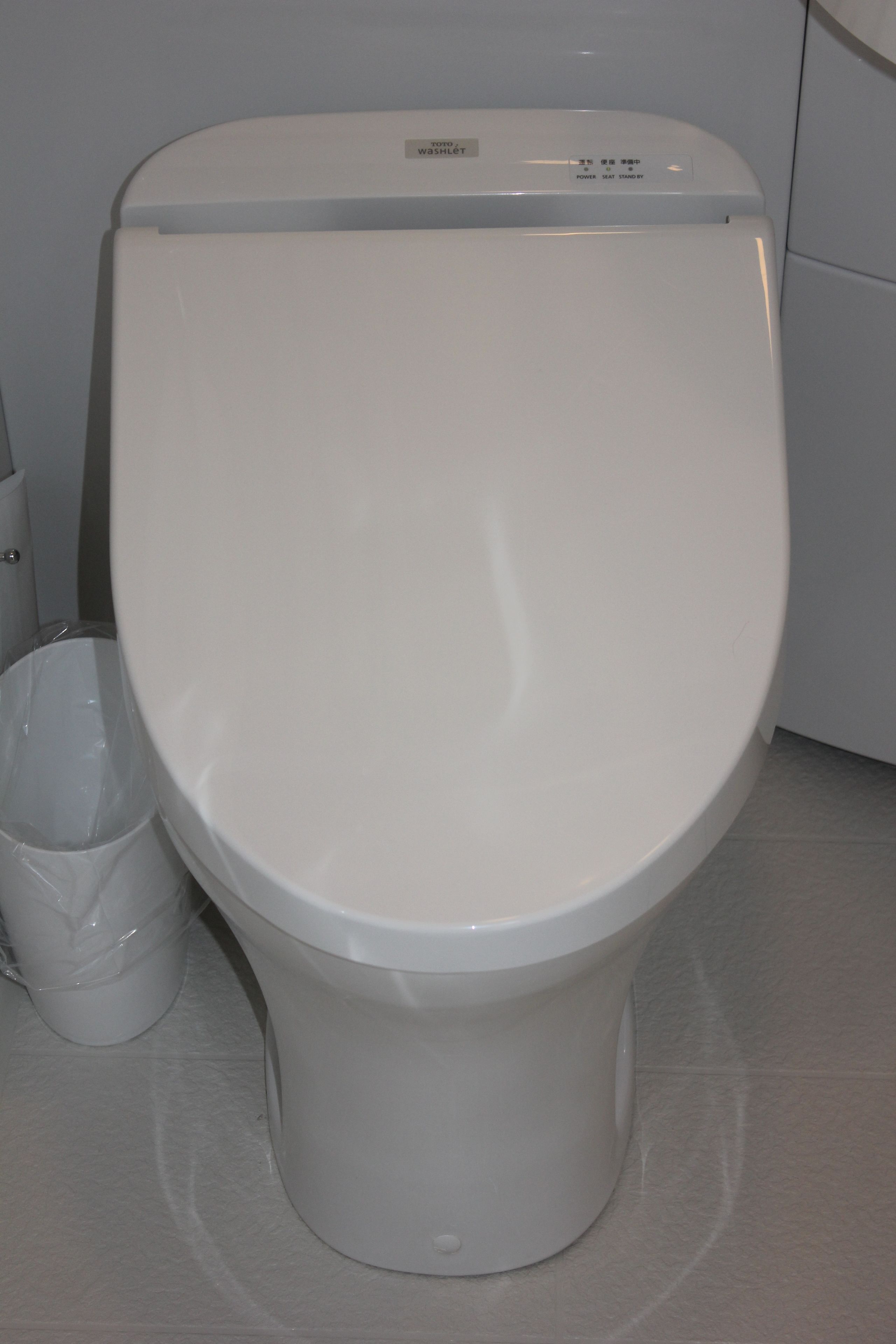 A white toilet with a control panel on top, a trash bin to the left, and a tiled floor.