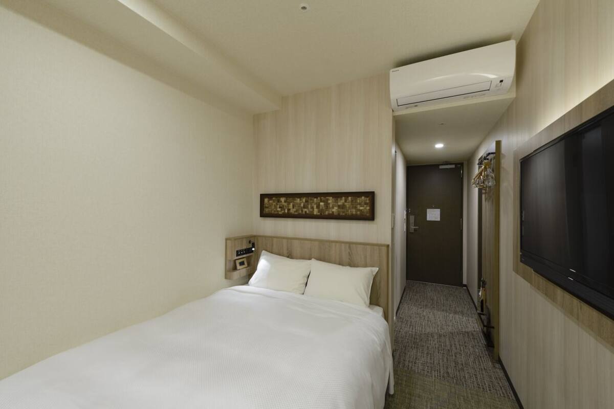 Quarto de hotel com uma cama, ar condicionado na parede, televisão e porta de acesso a outro cômodo.