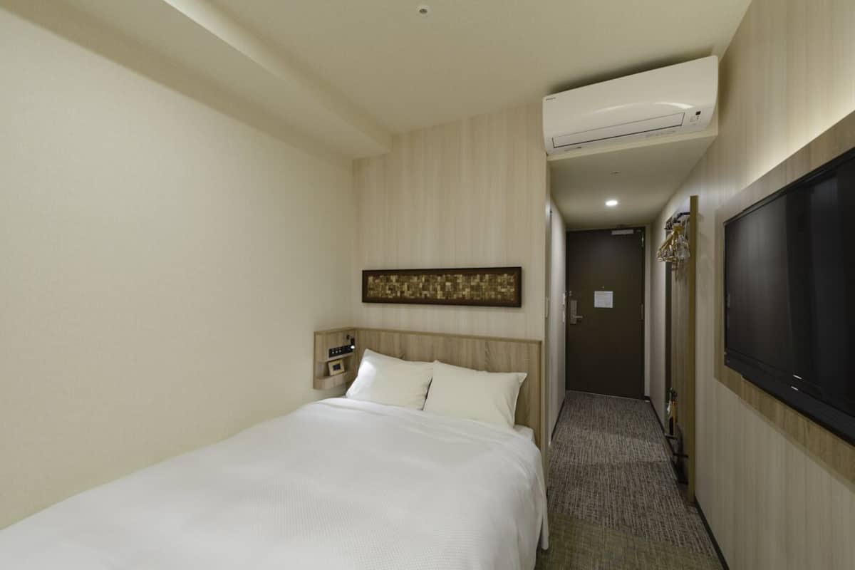 Habitación de hotel con cama, aire acondicionado instalado en la pared, televisor y puerta que da a otra habitación.