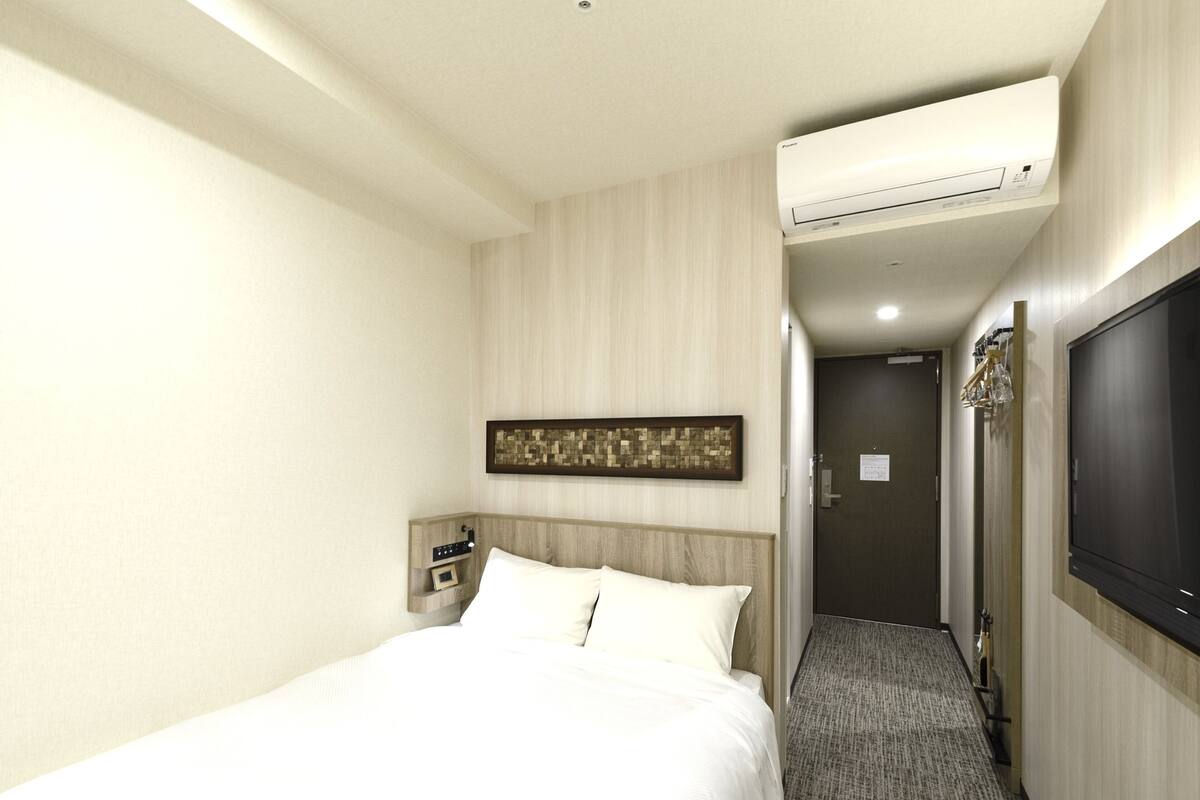 Quarto de hotel com uma cama, uma TV fixada na parede, aparelho de ar condicionado, uma obra de arte pendurada e uma porta que dá para outro quarto.