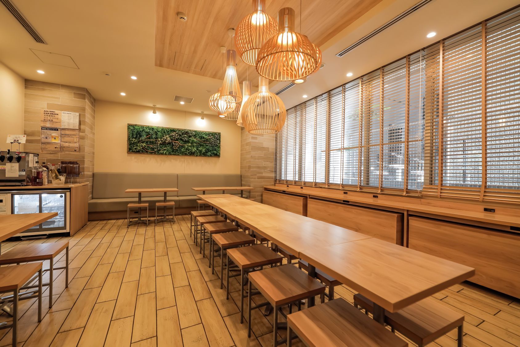 Interior de café com mesas e bancos de madeira, uma máquina de café e um grande janelão com persianas.