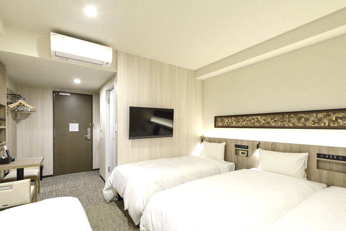 Quarto de hotel com duas camas, uma TV de tela plana na parede, uma mesa com cadeira e um porta-roupas.