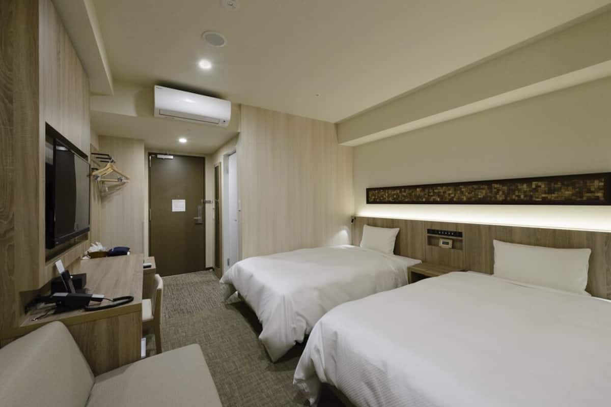 Quarto de hotel com duas camas, uma escrivaninha com cadeira, televisão e ar condicionado.