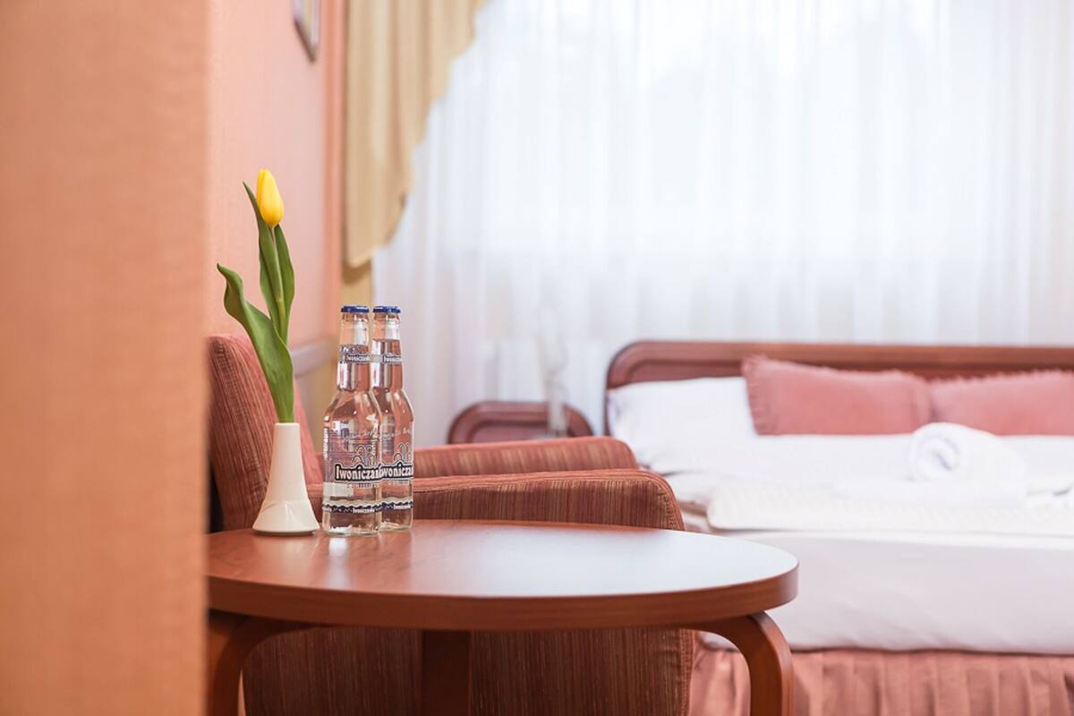 A hotel room with a bed, a bedside table with a bottle and a flower, and a window with curtains.