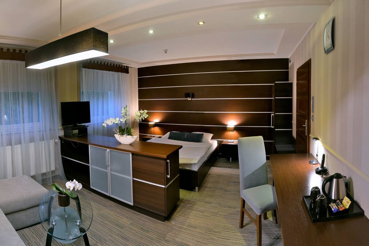 A modern hotel room with a sofa, a dining table, a TV, a bed, and a desk with a lamp.