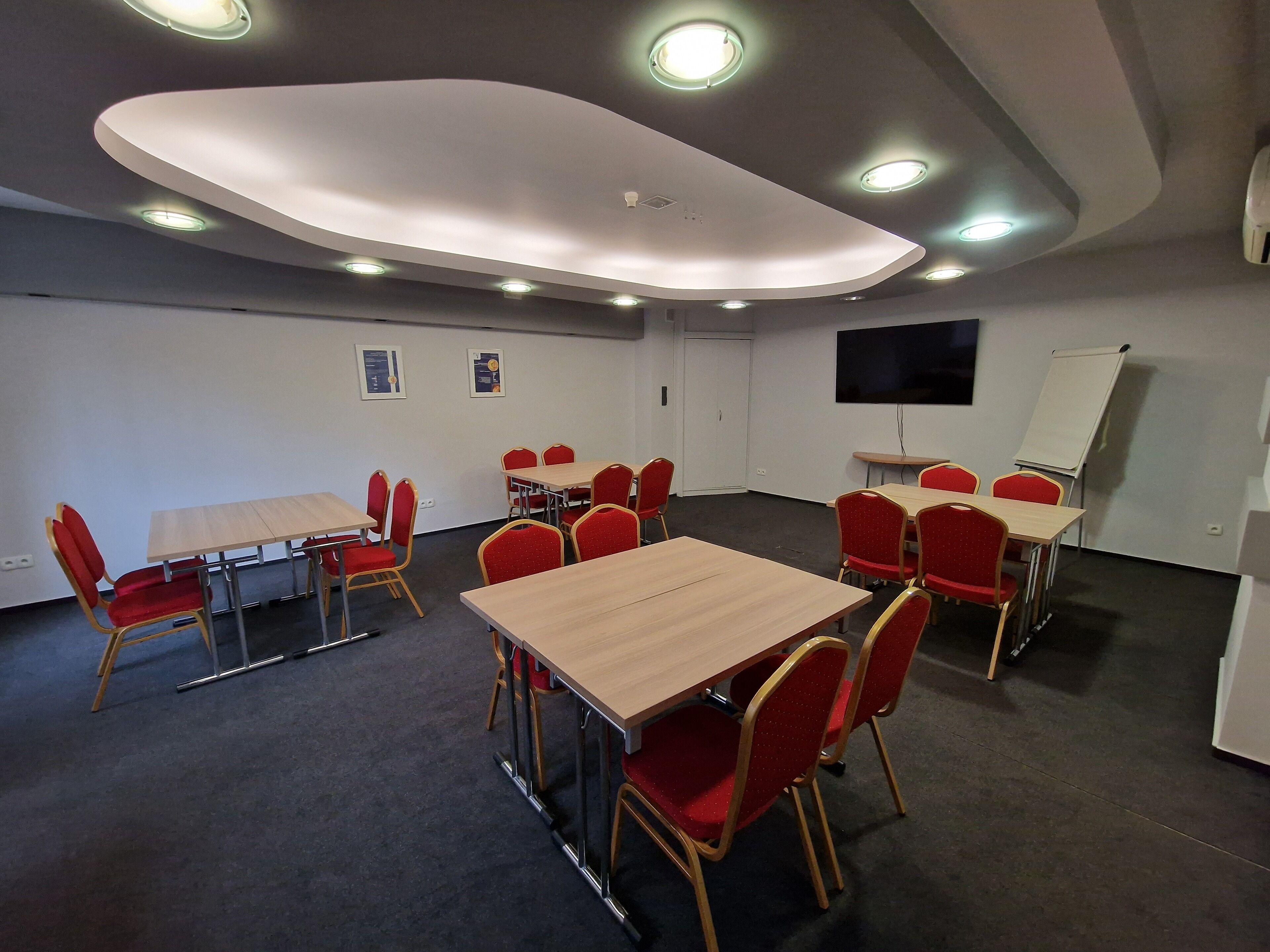 Une salle de conférence avec des tables rondes, des chaises rouges, un tableau blanc et un téléviseur à écran plat.