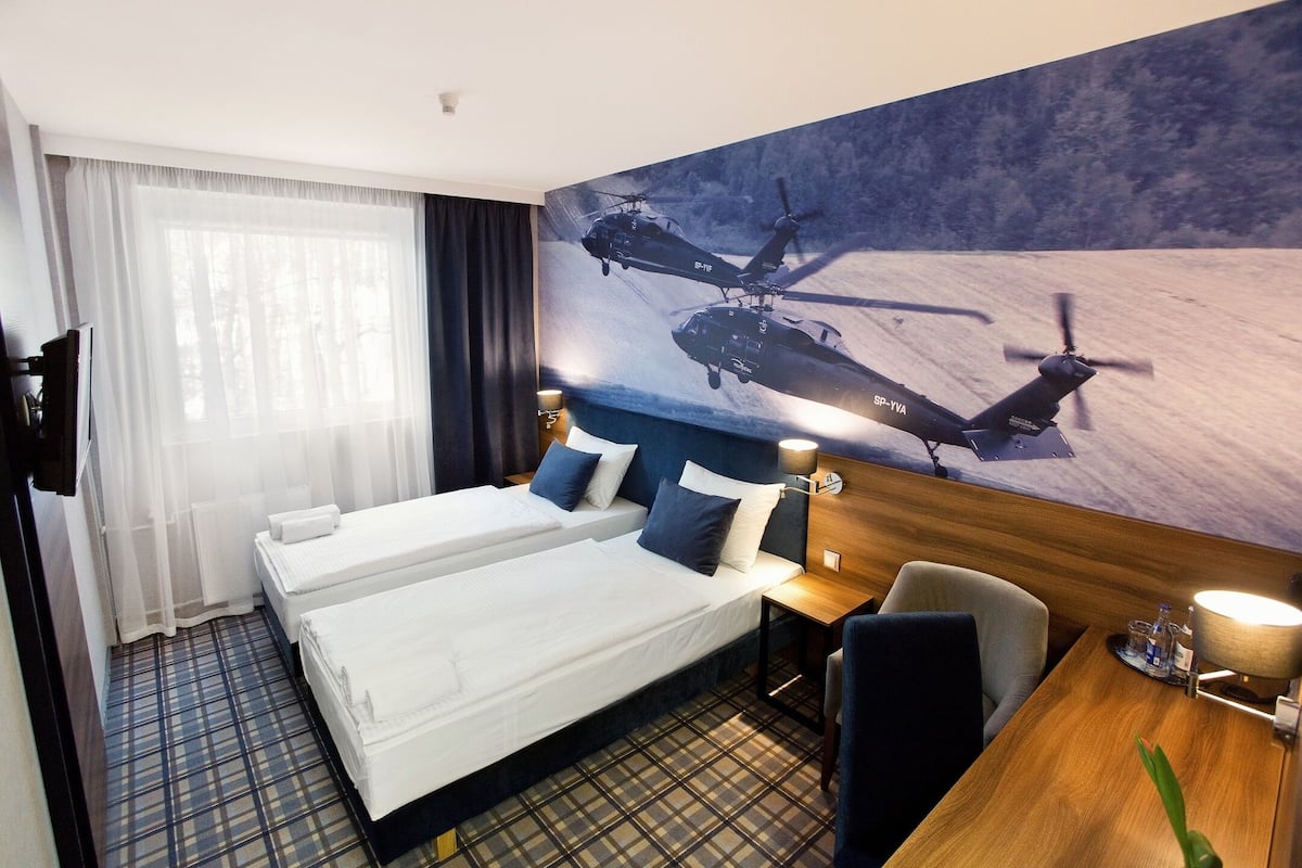 A hotel room with two beds, a large window with sheer curtains, a painting of helicopters, a wooden desk, and a chair.