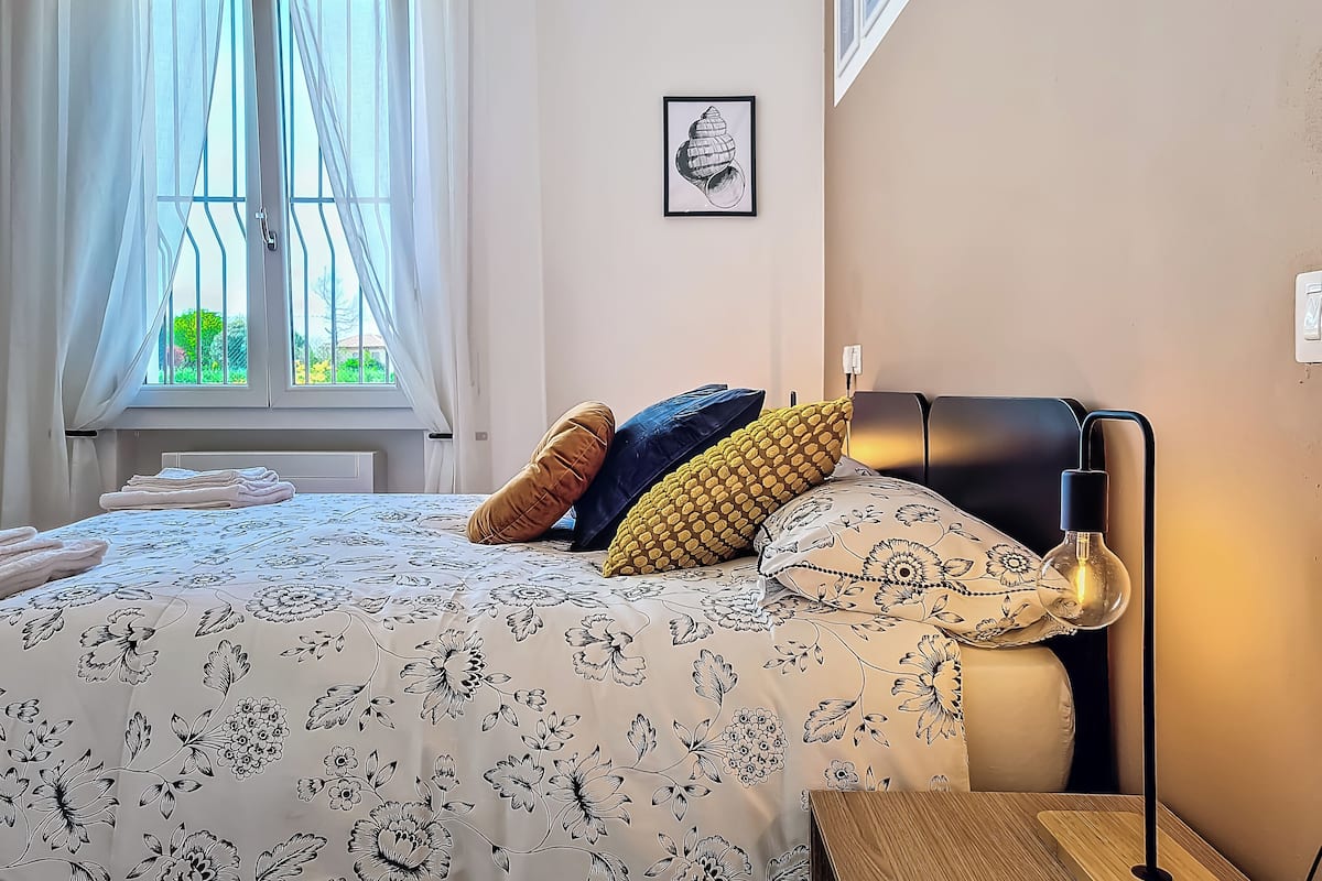 A neatly made bed with floral bedding, a bedside lamp, and a window with sheer curtains.
