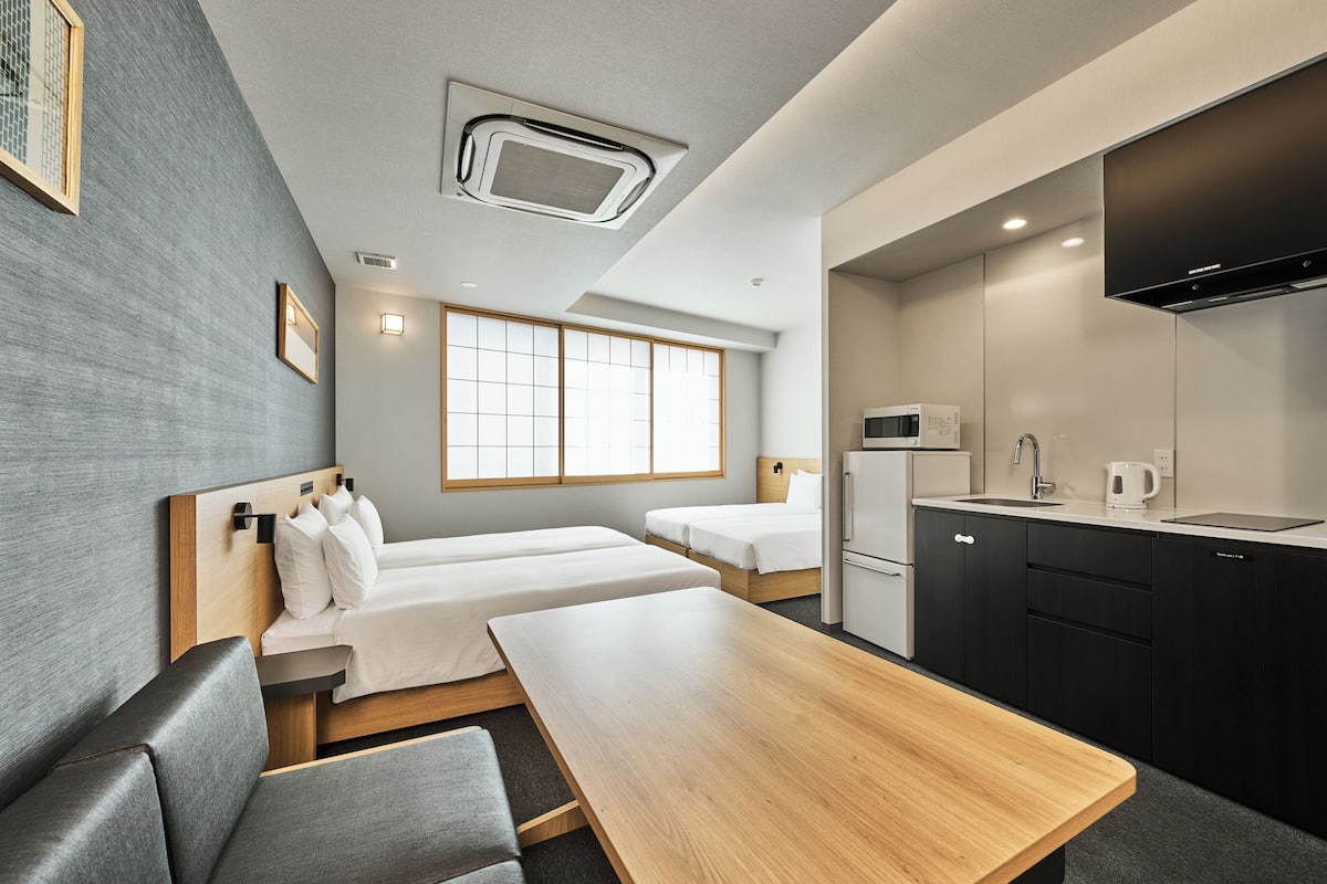 A modern hotel room with two beds, a wooden table, a kitchen area with a microwave, and a large window.