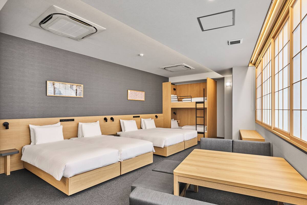 A modern hotel room with two single beds, a sofa, a table, and a bunk bed.